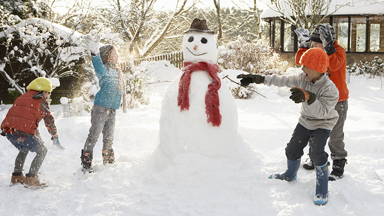 Mother And Children Building Snowman In Garden Having Snowball Fight