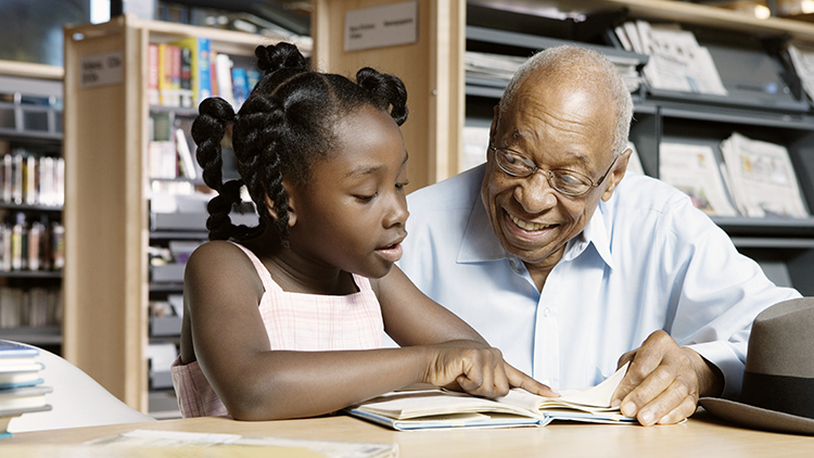 Seated view of a senior African-American man sitting with his granddaughter in a library while she reads a book aloud