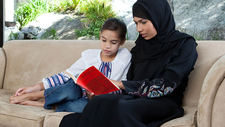 Arab mother and daughter reading book, sitting on sofa.