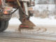 View from the car behind orange highway maintenance truck spreading deicing salt and sand, crystals dropping on the ice-covered asphalt road.