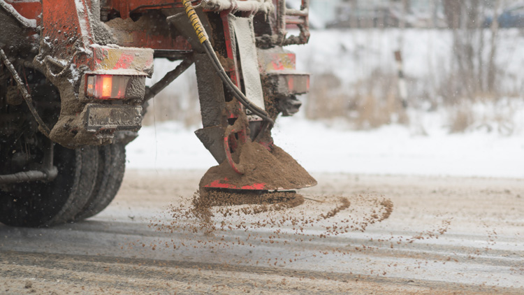 View from the car behind orange highway maintenance truck spreading deicing salt and sand, crystals dropping on the ice-covered asphalt road.
