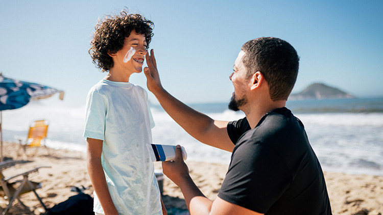 Papá aplicándole protector solar en la cara a su hijo sonriente en una playa soleada.