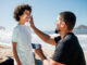 Dad applying sunscreen to a smiling son's face on a sunny beach.